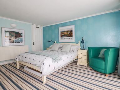 A bedroom with a bed a green armchair two framed pictures and a striped carpet at Blue Bay Beach House in Mawgan Porth