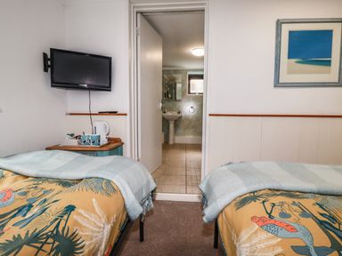 A bedroom with two beds a wall-mounted television a small table with a kettle mugs and an open door leading to a bathroom at Blue Bay Beach House in Mawgan Porth