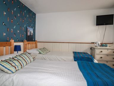 A bedroom with two single beds blue striped cushions and blankets a chest of drawers and a wall mounted television at Blue Bay Beach House in Mawgan Porth