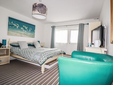 A bedroom with a bed a teal chair a television and a window with curtains at Blue Bay Beach House in Mawgan Porth