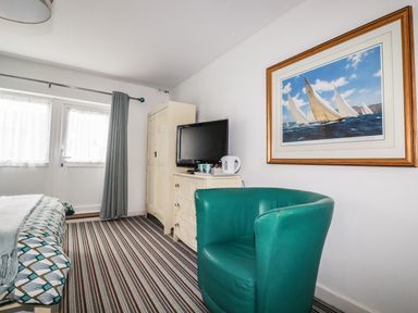 A room with a green armchair a dresser with a television and a kettle a cabinet and a bed with patterned bedding at Blue Bay Beach House in Mawgan Porth