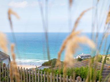 A view of the ocean with waves and a sandy beach behind a wooden fence and grass at Blue Bay Beach House in Mawgan Porth