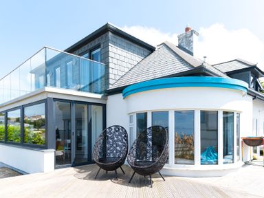 An outdoor patio with two black decorative chairs next to a modern house with a glass balcony and large windows at Blue Bay Beach House in Mawgan Porth