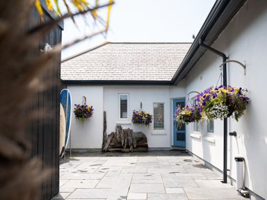 An outdoor courtyard with hanging flower baskets surfboards and wooden logs at Blue Bay Beach House in Mawgan Porth