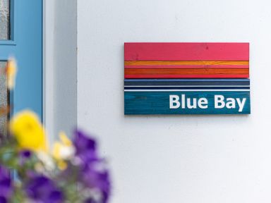 A colorful wooden sign reading Blue Bay on a white wall next to a blue door with flowers in the foreground at Blue Bay Beach House in Mawgan Porth