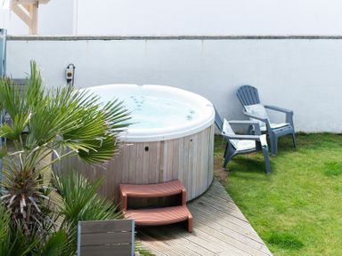 An outdoor area with a round hot tub wooden steps two plastic chairs with cushions and green grass at Blue Bay Beach House in Mawgan Porth