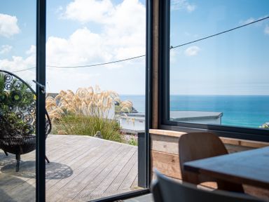 A dining area with wooden chairs and table next to a glass door opening to a deck with outdoor chair and a view of the sea at Blue Bay Beach House in Mawgan Porth
