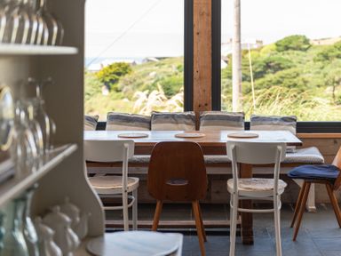 A dining area with a wooden table surrounded by chairs and a bench with cushions near a large window overlooking greenery at Blue Bay Beach House in Mawgan Porth