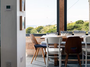 A dining area with a wooden table four chairs and a bench with cushions by a large window showing trees and hills at Blue Bay Beach House in Mawgan Porth