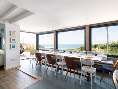 A dining area with a wooden table surrounded by chairs and large windows overlooking the sea at Blue Bay Beach House in Mawgan Porth