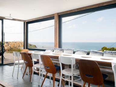 A dining room with a long wooden table chairs and large windows overlooking the sea at Blue Bay Beach House in Mawgan Porth