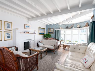 A living room with rattan furniture a white sofa a fireplace and large windows at Blue Bay Beach House in Mawgan Porth