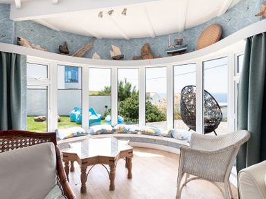 A curved seating area with cushions by large windows overlooking a garden with blue bean bags and a woven chair near a star-shaped wooden table at Blue Bay Beach House in Mawgan Porth