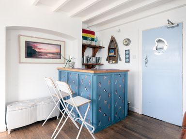 A small bar area with two white folding chairs a blue door and wall shelves at Blue Bay Beach House in Mawgan Porth