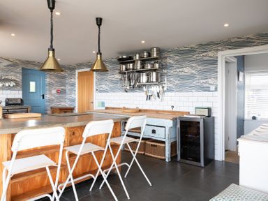 A kitchen with a wooden island and white chairs three pendant lights and utensils hanging on the wall at Blue Bay Beach House in Mawgan Porth