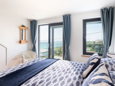 A bedroom with a bed facing windows with curtains overlooking the sea at Blue Bay Beach House in Mawgan Porth