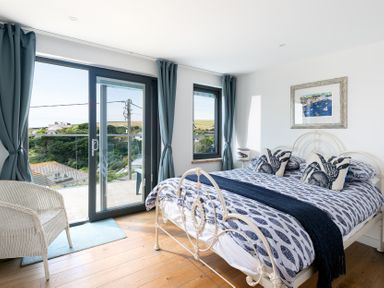 A bedroom with a double bed blue curtains wicker chair and glass doors opening to a balcony at Blue Bay Beach House in Mawgan Porth