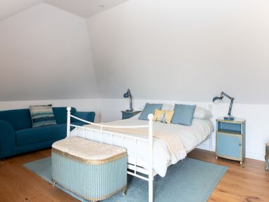A bedroom with a white bed, blue sofa, blue rug, and bedside tables with lamps at Blue Bay Beach House in Mawgan Porth