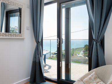 A room with large glass doors showing a sea view with blue curtains and a wicker chair with a pillow at Blue Bay Beach House in Mawgan Porth