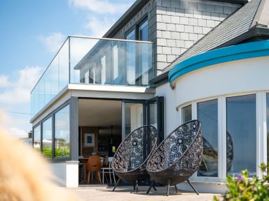 A patio area with two patterned chairs in front of a house with glass balcony and large windows at Blue Bay Beach House in Mawgan Porth