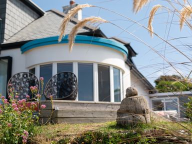 A garden with a wooden sculpture pink flowers and patio chairs outside a house with curved windows at Blue Bay Beach House in Mawgan Porth