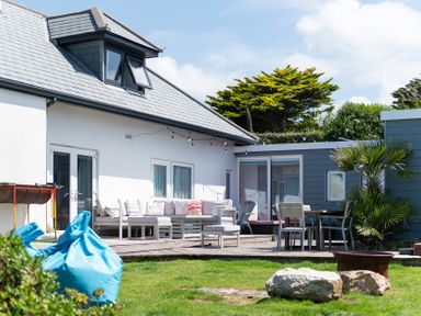 An outdoor patio area with seating furniture including a sofa and chairs on a wooden deck next to a white and blue house with a garden in the foreground at Blue Bay Beach House in Mawgan Porth