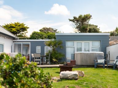 A backyard with a wooden deck dining table set a hot tub lawn chairs fire pit and shrubs at Blue Bay Beach House in Mawgan Porth