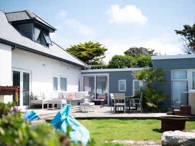 An outdoor patio area with seating and dining furniture next to a white and blue house surrounded by greenery at Blue Bay Beach House in Mawgan Porth
