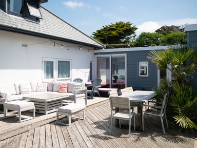 An outdoor wooden deck with gray seating furniture a table chairs string lights and plants at Blue Bay Beach House in Mawgan Porth