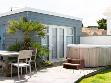 An outdoor deck area with a table and chairs next to a small building with blue siding and glass doors with a hot tub at Blue Bay Beach House in Mawgan Porth