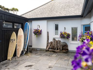 An outdoor area with three surfboards leaning against a black wall hanging flower baskets and pieces of driftwood at Blue Bay Beach House in Mawgan Porth