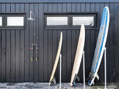 Three surfboards standing on racks next to an outdoor shower mounted on a black wooden wall with two small windows at Blue Bay Beach House in Mawgan Porth