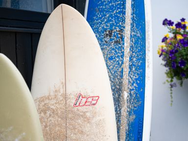 Three surfboards leaning against a black wall near a white wall with a hanging flower planter with purple and yellow flowers at Blue Bay Beach House in Mawgan Porth