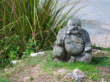 A stone statue of a laughing Buddha sitting on grass with plants in the background at Blue Bay Beach House in Mawgan Porth