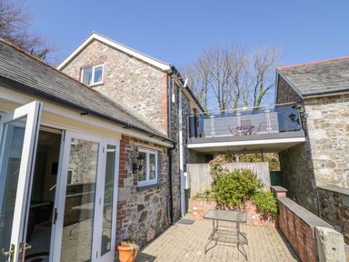 A stone building with a glass door opening to a patio with a small table and a balcony with outdoor furniture at Lambkin Cottage in St Columb Major