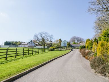 A paved driveway with a fence and grass on the left and bushes on the right leading to multiple houses in the background at Lambkin Cottage in St Columb Major