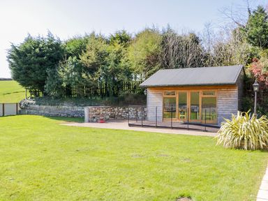 A wooden outbuilding with glass doors on a paved area surrounded by grass and trees at Lambkin Cottage in St Columb Major