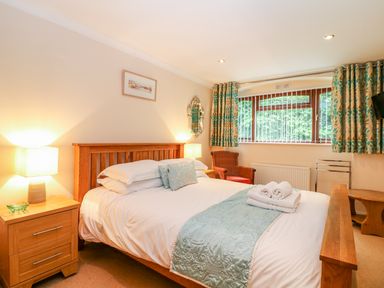 A bedroom with a wooden bed with white bedding two bedside tables with lamps a wicker chair near a window with patterned curtains at Lambkin Cottage in St Columb Major