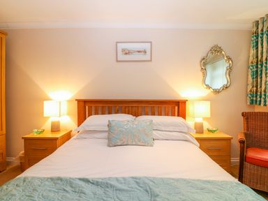 A bedroom with a double bed lamps on wooden nightstands a wicker chair and a decorative mirror at Lambkin Cottage in St Columb Major