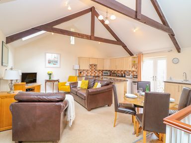 An open plan living room with brown leather sofas a yellow chair a wooden dining table with four chairs a kitchen area with light wood cabinets and tiled backsplash at Lambkin Cottage in St Columb Major
