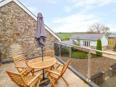 A wooden patio table with umbrella and chairs on a balcony overlooking a courtyard with green grass and buildings at Lambkin Cottage in St Columb Major