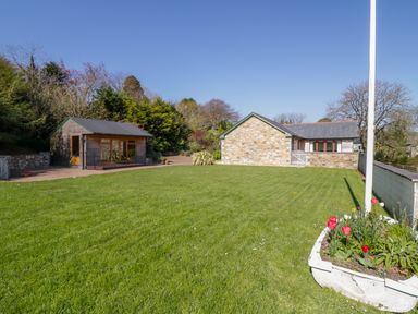A garden with a green lawn red flowers a wooden shed and a stone building at Lambkin Cottage in St Columb Major