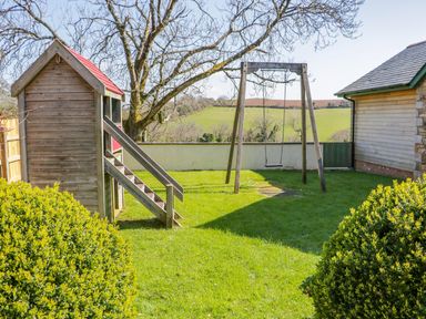A backyard with a wooden playhouse and a swing set surrounded by grass and bushes at Lambkin Cottage in St Columb Major