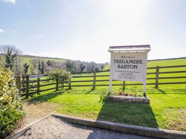 A wooden sign welcoming to Tregamere Barton with a gravel path and wooden fence in a green rural landscape at Lambkin Cottage in St Columb Major