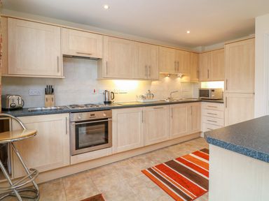 A kitchen with wooden cabinets, electric oven, kettle, toaster, microwave, sink, and striped rugs on the floor at Cornflower Cottage in St Columb Major