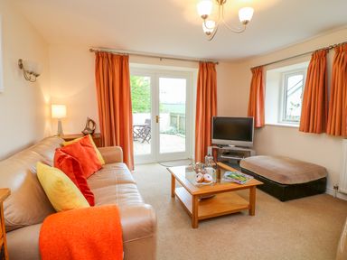 A living room with a beige leather sofa, orange curtains, a wooden coffee table, television, and patio doors at Cornflower Cottage in St Columb Major