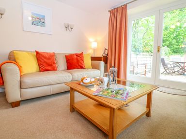A living room with a beige sofa with orange and yellow cushions and a wooden coffee table with magazines and a coffee set at Cornflower Cottage in St Columb Major