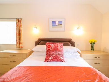A bedroom with a bed with orange bedding and a red pillow between two wooden nightstands with flowers and a window at Cornflower Cottage in St Columb Major