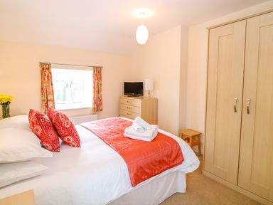 A bedroom with a bed with red pillows and orange runner a wooden wardrobe and dresser with television at Cornflower Cottage in St Columb Major