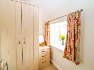 A bedroom corner with a wooden wardrobe and dresser near a window with patterned curtains and a vase of flowers at Cornflower Cottage in St Columb Major